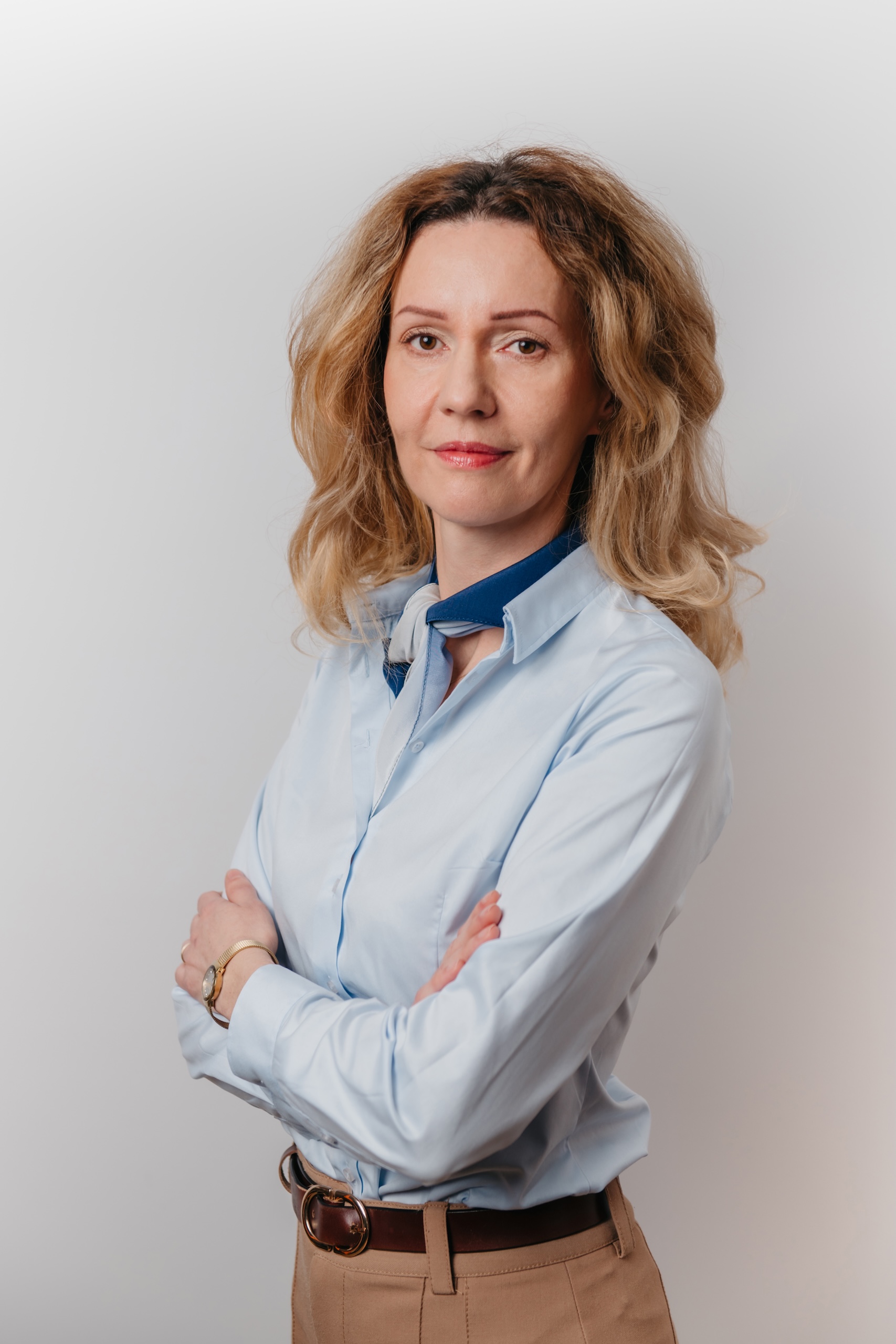Kobieta z falistymi blond włosami ubrana w jasnoniebieską koszulę zapinaną na guziki i beżowe spodnie stoi ze skrzyżowanymi ramionami na jasnym tle.