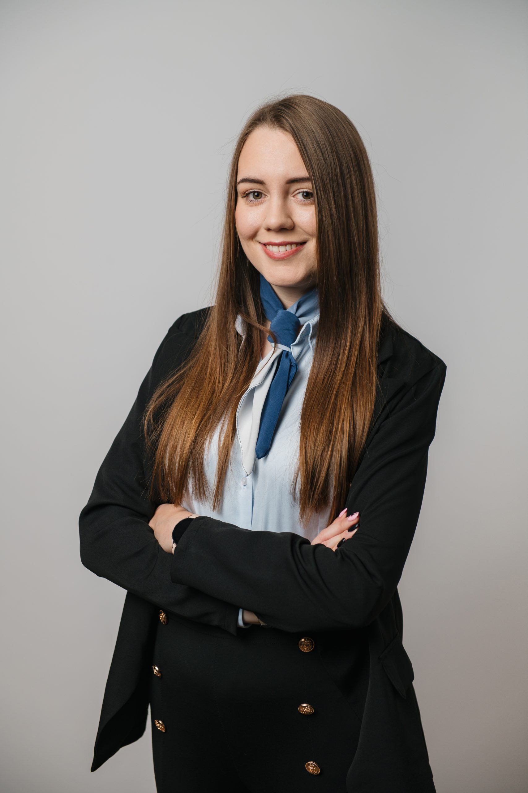 Młoda kobieta z długimi prostymi włosami stoi na prostym tle, ubrana w czarną marynarkę i jasnoniebieską bluzkę z niebieskim krawatem, ze skrzyżowanymi ramionami i uśmiechem.