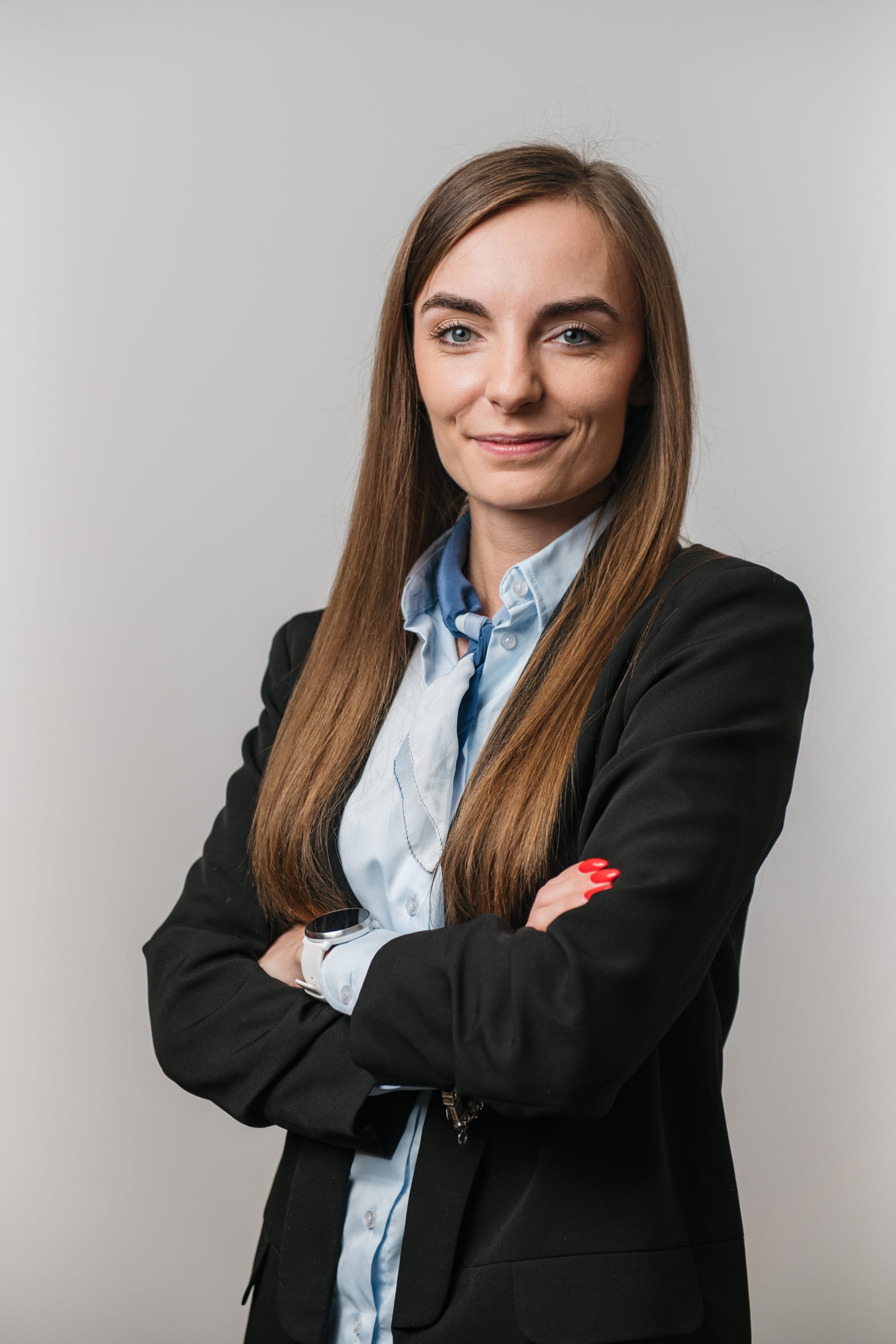 Kobieta z długimi prostymi włosami, ubrana w czarną marynarkę i jasnoniebieską koszulę, stoi ze skrzyżowanymi ramionami, patrząc pewnie w kamerę na prostym tle.