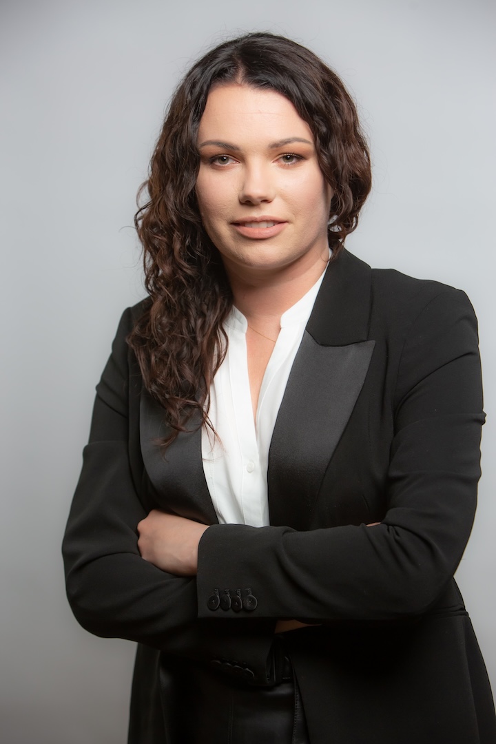 Kobieta z długimi kręconymi brązowymi włosami ubrana w czarną marynarkę i białą bluzkę stoi ze skrzyżowanymi ramionami na prostym tle.