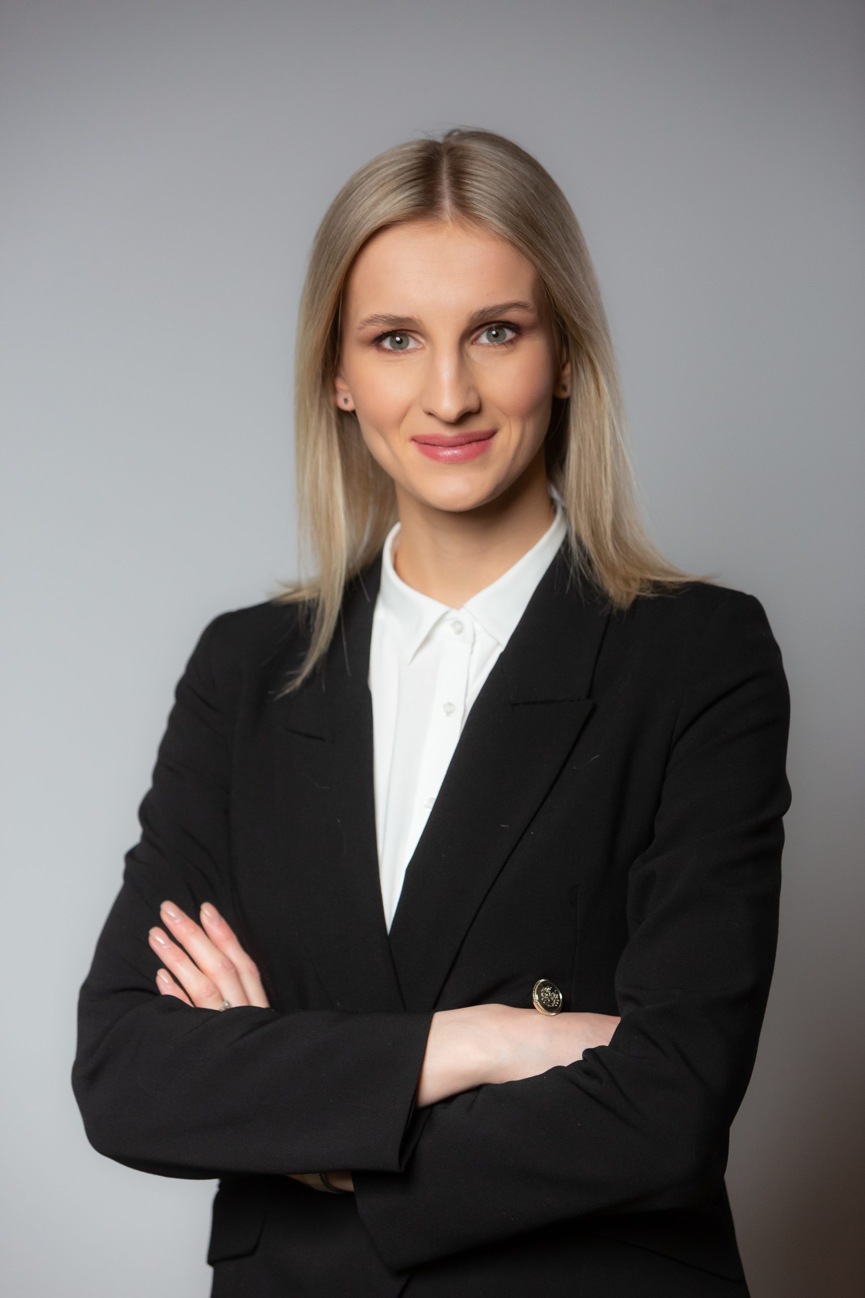 Kobieta o prostych blond włosach ubrana w czarną marynarkę i białą koszulę stoi ze skrzyżowanymi ramionami na jasnym tle.