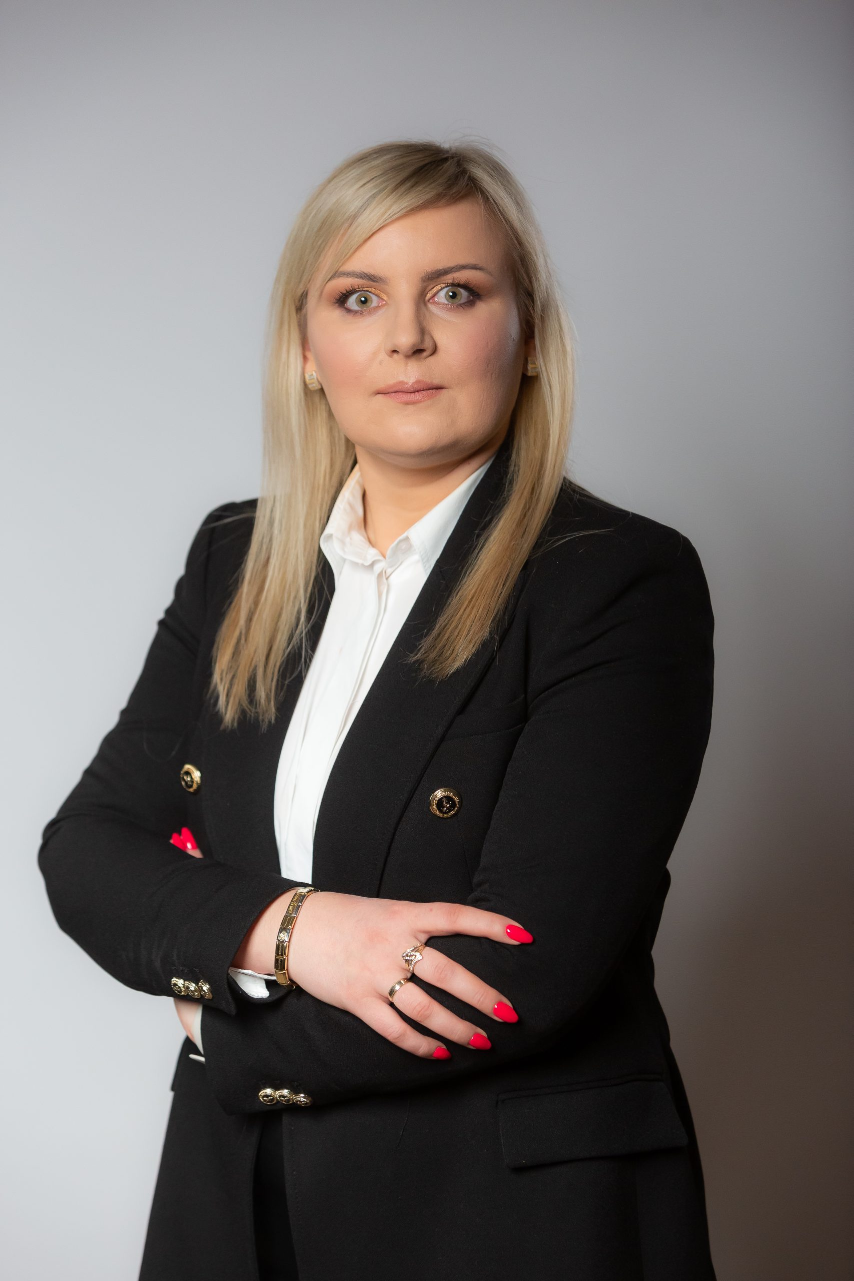 Kobieta o prostych blond włosach, ubrana w czarną marynarkę i białą koszulę, stoi ze skrzyżowanymi ramionami na prostym tle.