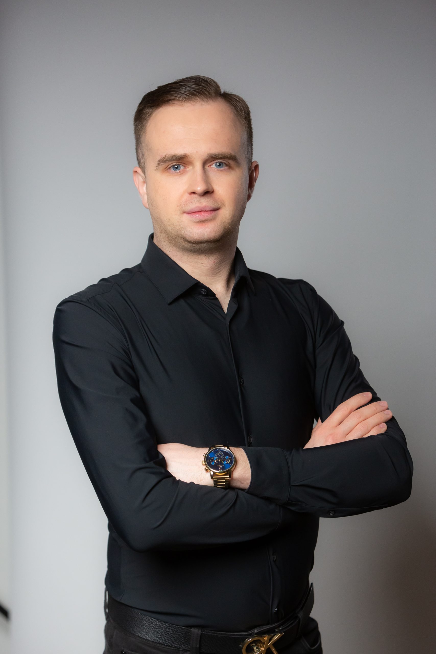 Mężczyzna z krótkimi włosami ubrany w czarną koszulę zapinaną na guziki i zegarek stoi ze skrzyżowanymi rękami na prostym tle.