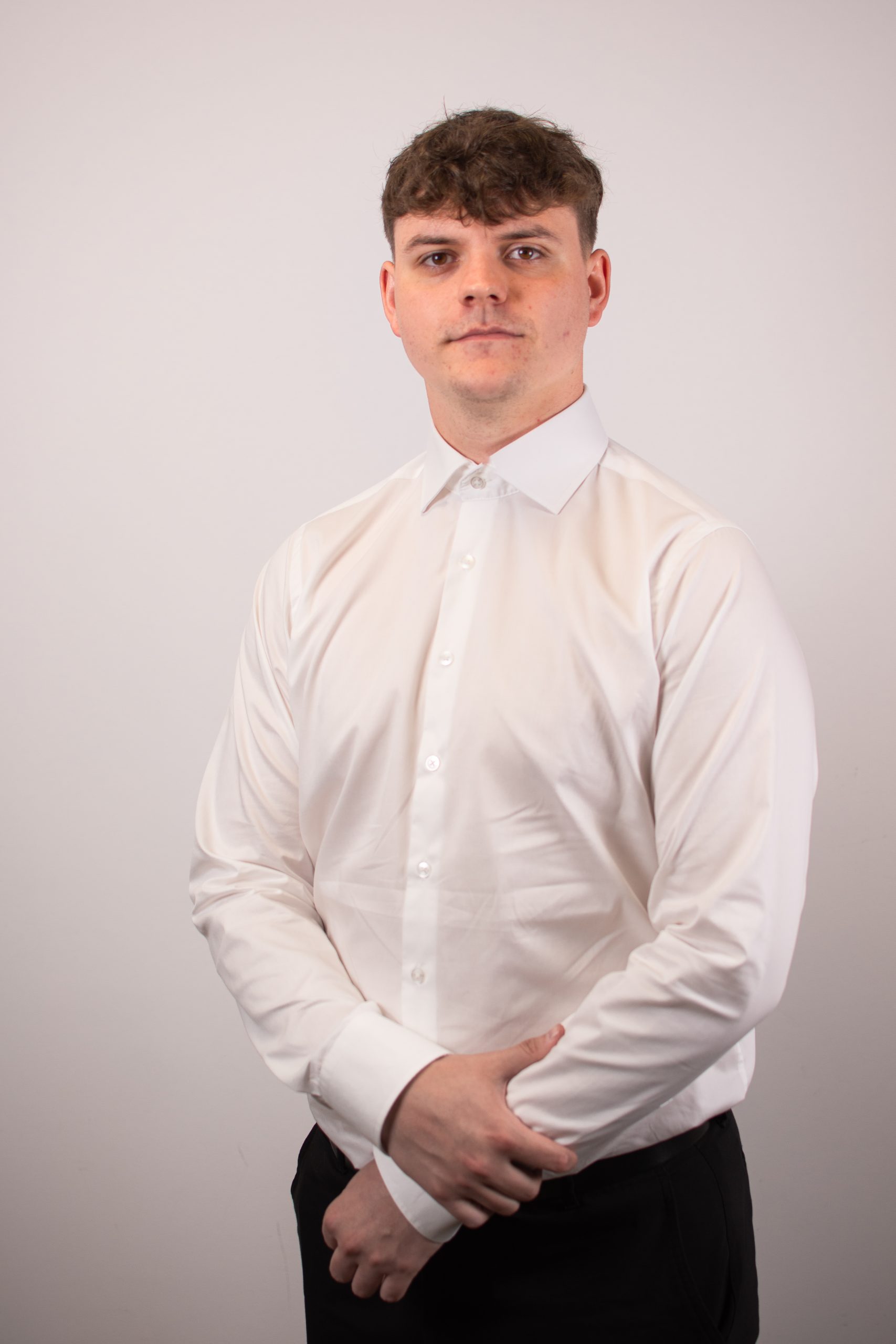 Młody mężczyzna z krótkimi brązowymi włosami stoi na jasnym tle, ubrany w białą koszulę zapinaną na guziki i ciemne spodnie, z rękami splecionymi z przodu.