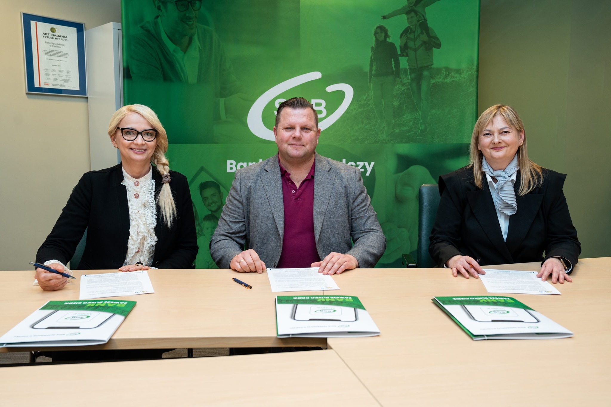 Trzy osoby w strojach biznesowych siedzą przy stole z dokumentami i długopisami, pozując do zdjęcia przed zielonym banerem z logo.