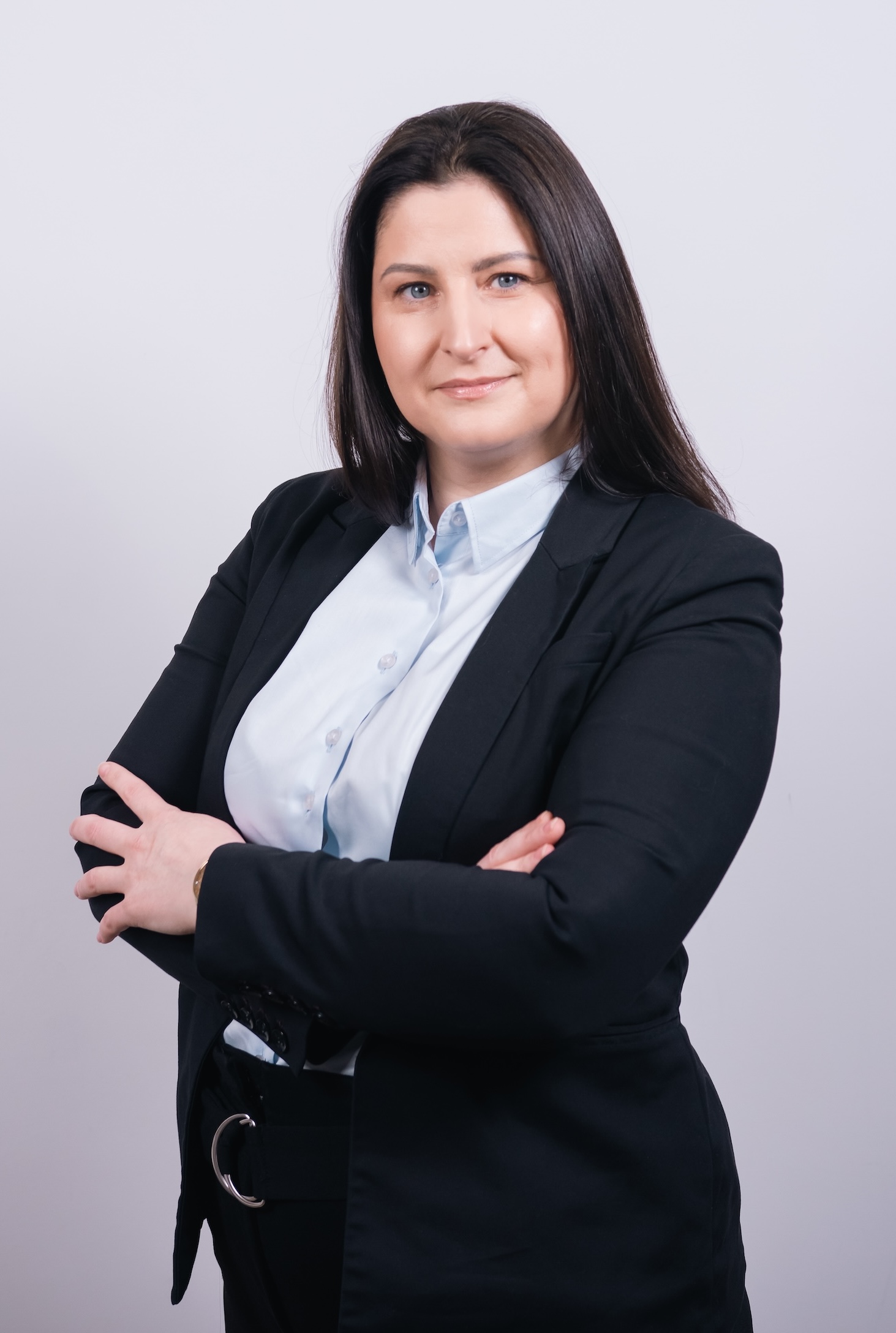 Kobieta z długimi ciemnymi włosami w czarnym garniturze i jasnoniebieskiej koszuli stoi ze skrzyżowanymi ramionami na prostym, jasnym tle.